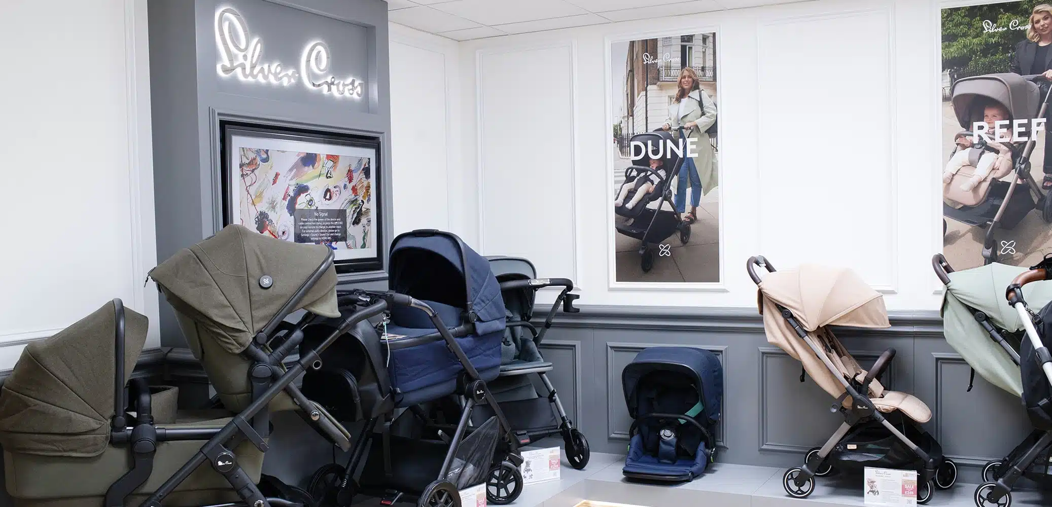 Display area for baby strollers with illuminated "Silver Cross" sign and "DUNE" and "Reef" pushchair posters.